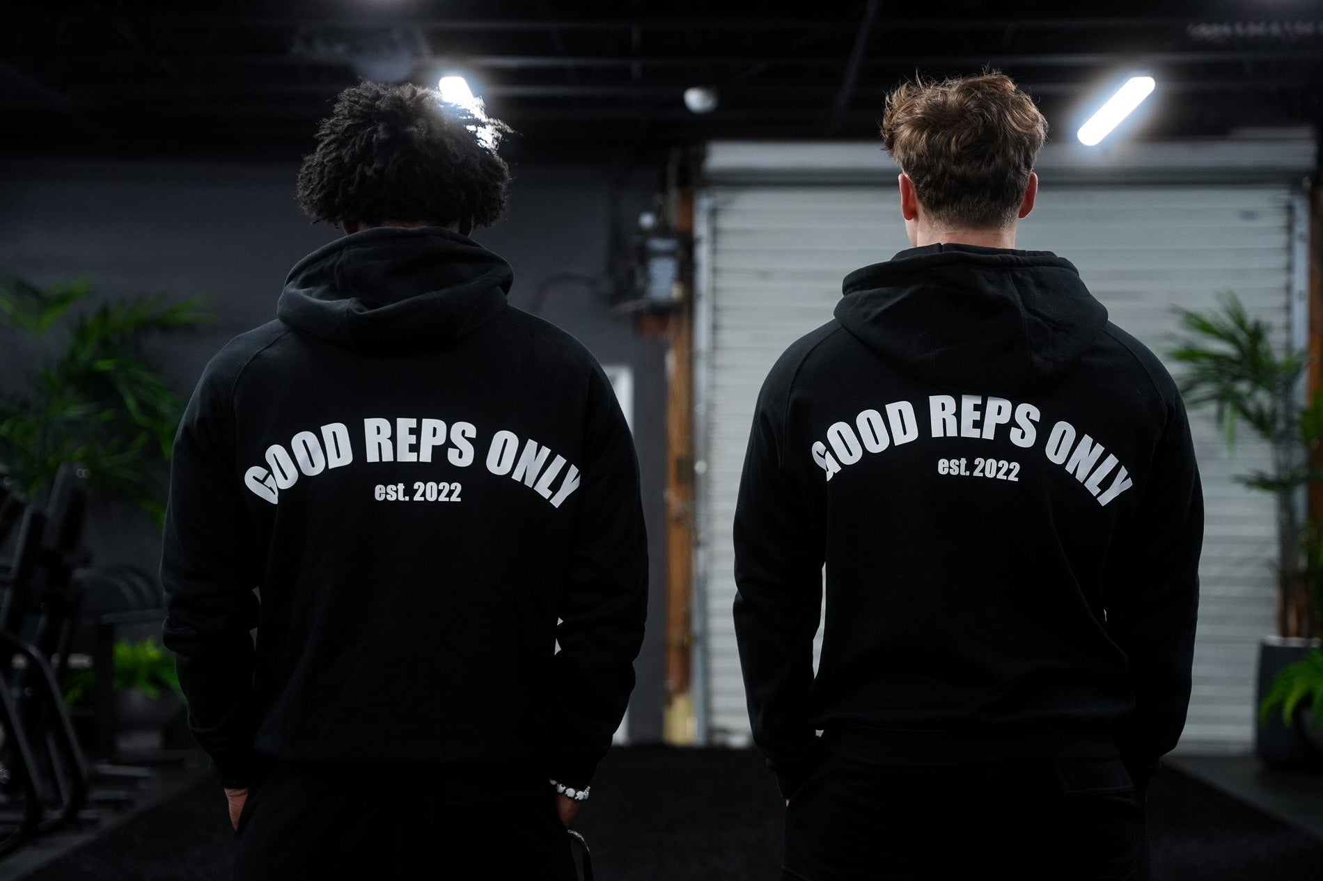 Two people wearing black hoodies with 'Good Reps Only' text in a dark indoor setting.
